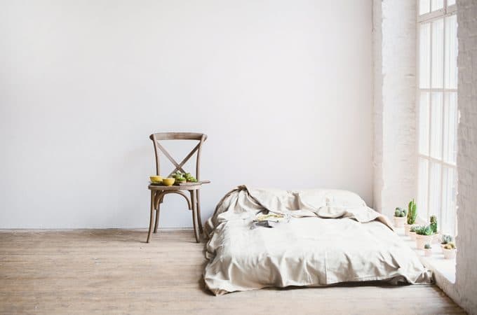 A minimalist bedroom with a basic bed frame, a plain white duvet, and bare walls. There's a simple rug on the floor and a single, understated nightstand.