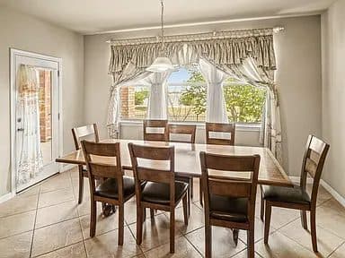 A cluttered, dated dining room with a heavy wooden table, mismatched chairs, and dark decor elements. The walls are a neutral beige, and the window has simple, dark curtains.