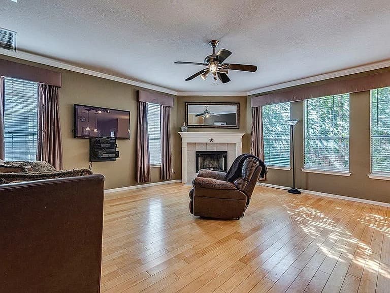 A dated living room with olive-brown walls, satin valances over the windows, a brown leather recliner sitting mid-room, a wall-mounted TV, and a corner fireplace with a mirror above it.