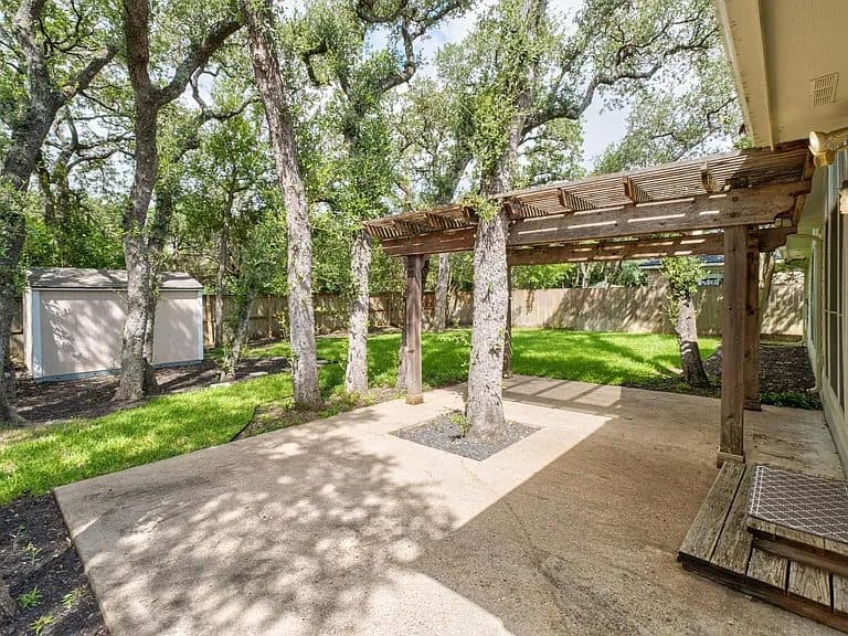 Before: an empty concrete patio under a weathered wood pergola, with large live oak trees, a lawn, a wood fence, a garden shed and a small wooden step by the back door