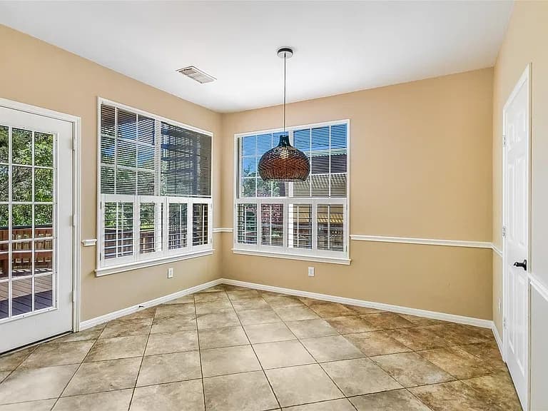 A bright, empty breakfast nook with tan walls, a white chair rail, tile floors, a glass door to the deck, large windows with half-raised blinds, and a woven rattan pendant light.