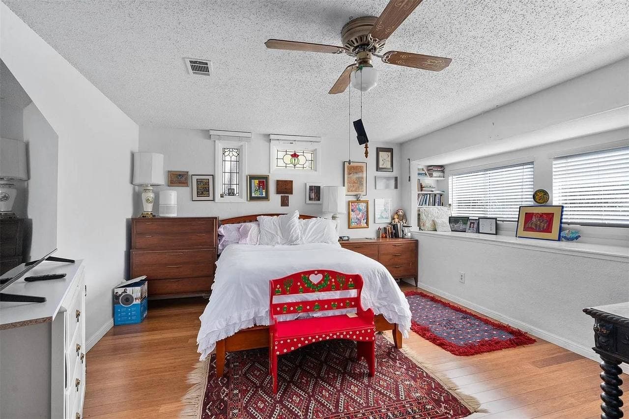 A lived-in bedroom with a queen bed in white ruffled bedding, a bright red painted bench, layered red oriental rugs, two mismatched wood dressers, stained-glass windows, and a gallery wall of small frames.