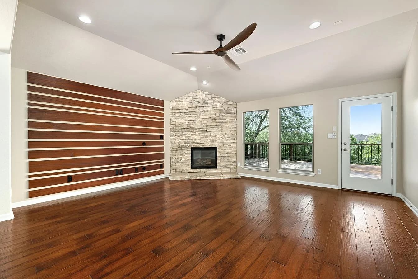 Before: an empty living room with dark hardwood floors, a horizontal wood-slat accent wall, a corner stacked-stone fireplace, two windows and a glass door to a deck
